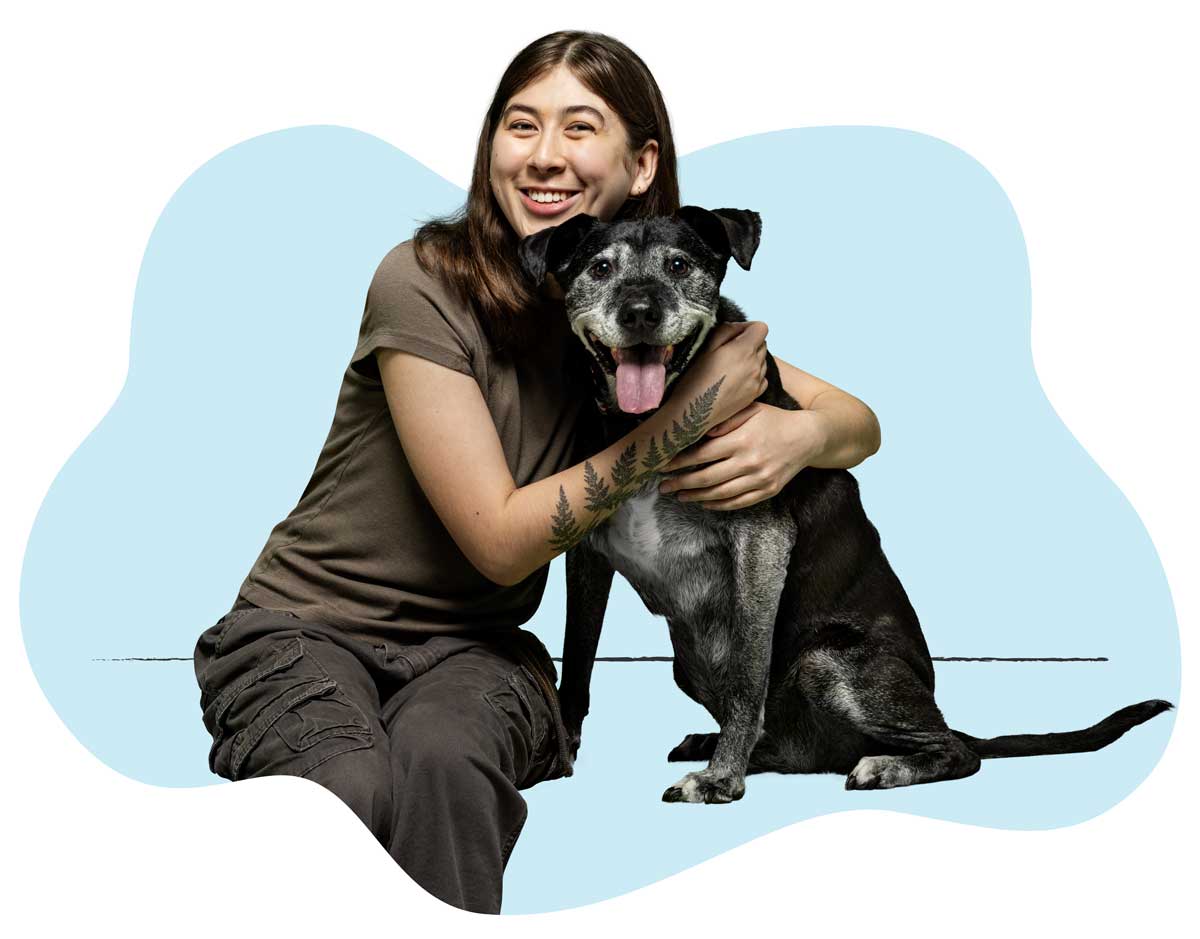 A woman in a brown shirt sitting on the floor hugging her black-haired dog